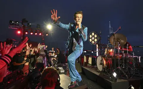 Brandon Flowers de The Killers, en costume bleu scintillant, salue la foule lors d'un concert. Les albums de The Killers résonnent dans la nuit.