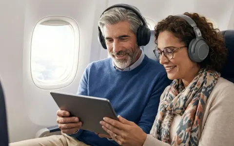 Couple souriant dans un avion, partageant une tablette et des écouteurs. Apprenez comment connecter deux appareils Bluetooth en même temps pour un voyage agréable.