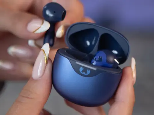 Une main aux ongles soignés tient un écouteur bleu qui semble trop grand pour l'oreille. L'étui de chargement est ouvert, révélant un autre écouteur à l'intérieur.
