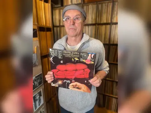 Un homme tient la pochette de l'album "One Size Fits All" de Frank Zappa, entouré de vinyles.