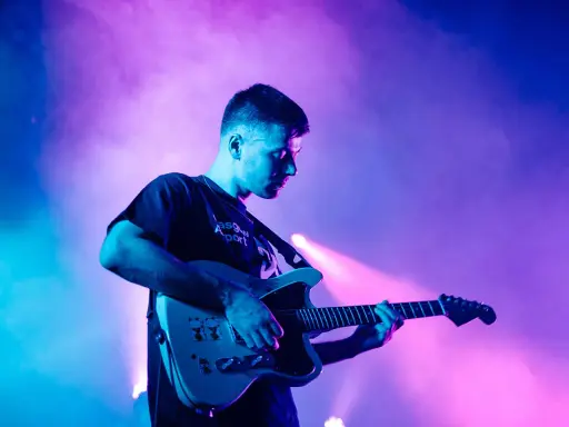 Un jeune homme joue de la guitare électrique sous des lumières bleues et roses. Il est en train de trouver des musiciens pour son groupe.