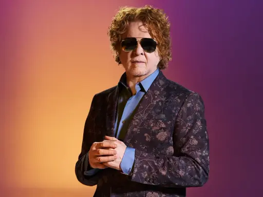 Homme aux cheveux bouclés roux, portant des lunettes de soleil et un costume fleuri, sur fond dégradé orange et violet. Il ressemble à un i monster.