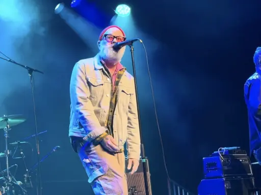 Le chanteur principal du rem groupe, avec une barbe blanche et un bonnet rouge, chante sur scène. Un guitariste est à droite.