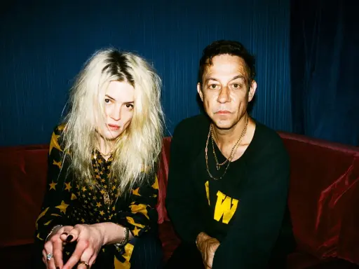 Un couple, aux allures rock, pose sur un canapé rouge. Elle, aux cheveux blonds décoiffés, porte une robe à étoiles. Lui, au regard intense, arbore un sweat noir. Ils semblent vouloir "kill the young" par leur style.