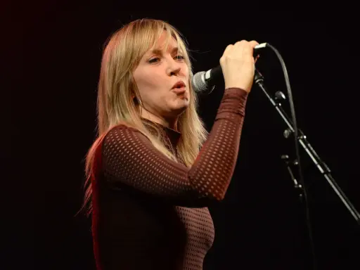 Liz Phair chante avec passion dans un micro, ses cheveux blonds encadrant son visage expressif.
