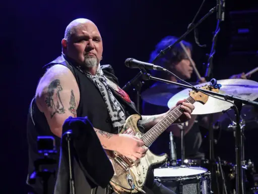 Un guitariste popa chubby, chauve, tatoué, joue sur scène avec un batteur en arrière-plan.