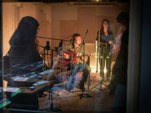 Un homme joue de la guitare dans un studio d'enregistrement, entouré de microphones. Une femme chante à ses côtés. La scène suggère une collaboration musicale, peut-être pour une liste producteur de musique.