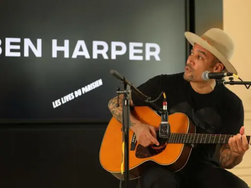 Ben Harper, chapeau sur la tête, joue de la guitare devant un écran affichant son nom et "Les Vies du Parisien".