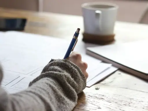 Main tenant une plume bleue, une personne écrit sur du papier, une tasse de café à proximité. Une idée de chanson à écrire prend forme.