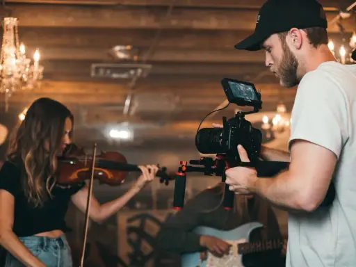 Un caméraman s'apprête à créer un clip vidéo, filmant une violoniste et un guitariste dans un décor chaleureux avec des lustres.
