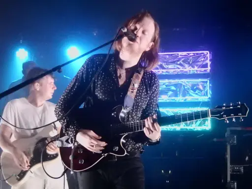 Le groupe Two Doors Cinema Club en concert, éclairé par des néons bleus. Le chanteur joue de la guitare et chante dans un micro.
