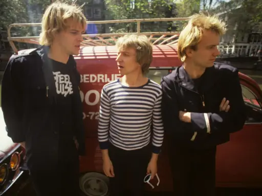 Le groupe Police, dans les années 80, pose devant un véhicule rouge. L'un porte un t-shirt à rayures, les autres des vestes sombres.