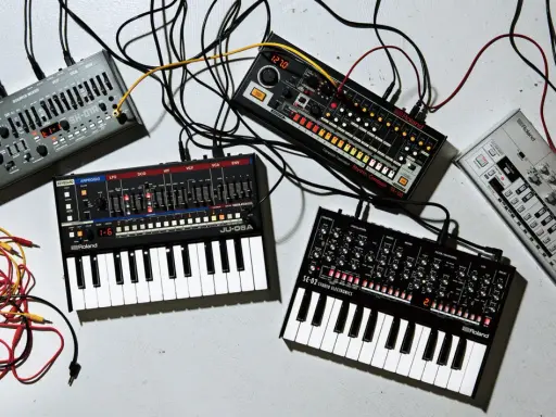 Plusieurs petits instruments de musique électroniques, dont des synthétiseurs et des boîtes à rythmes, sont connectés par des câbles.