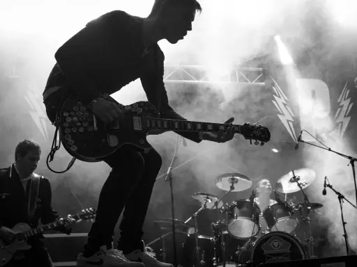 Un guitariste électrique joue sur scène, entouré de fumée. Derrière lui, un batteur et le logo d'un nom de groupe de rock.