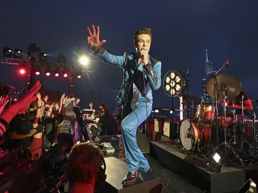 Brandon Flowers de The Killers, en costume bleu scintillant, salue la foule lors d'un concert. Les albums de The Killers résonnent dans la nuit.