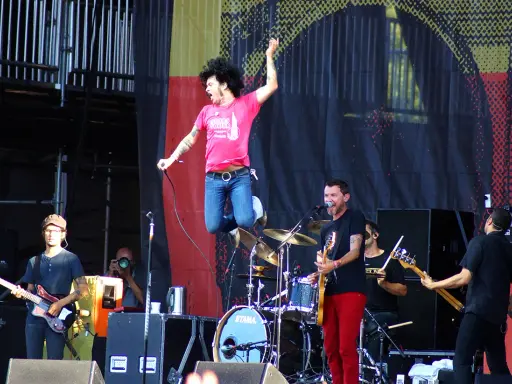 Un chanteur saute sur scène, micro en main, lors d'un concert en plein air. Le groupe joue avec énergie, une ambiance de festival.