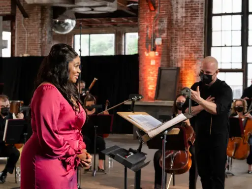 Une chanteuse en robe rose discute avec un chef d'orchestre masqué, entourée de musiciens. Elle cherche peut-être à trouver un agent pour sa carrière.
