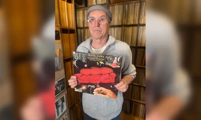 Un homme tient la pochette de l'album "One Size Fits All" de Frank Zappa, entouré de vinyles.
