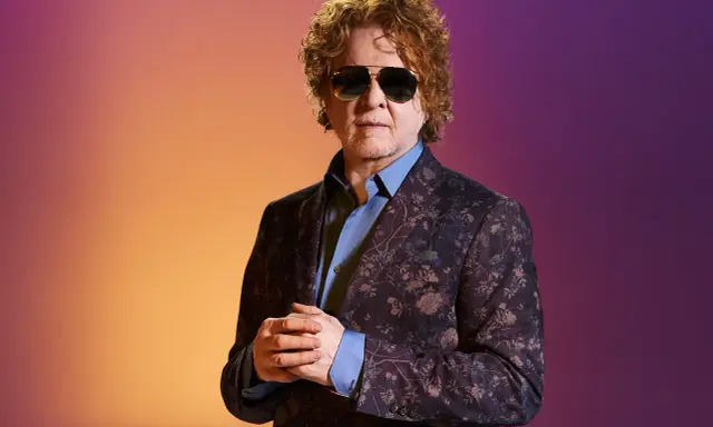Homme aux cheveux bouclés roux, portant des lunettes de soleil et un costume fleuri, sur fond dégradé orange et violet. Il ressemble à un i monster.