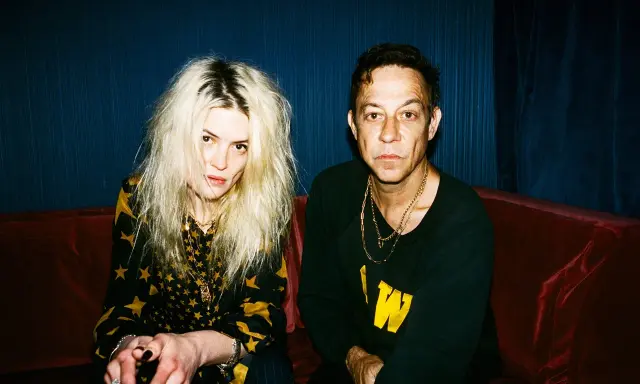 Un couple, aux allures rock, pose sur un canapé rouge. Elle, aux cheveux blonds décoiffés, porte une robe à étoiles. Lui, au regard intense, arbore un sweat noir. Ils semblent vouloir "kill the young" par leur style.