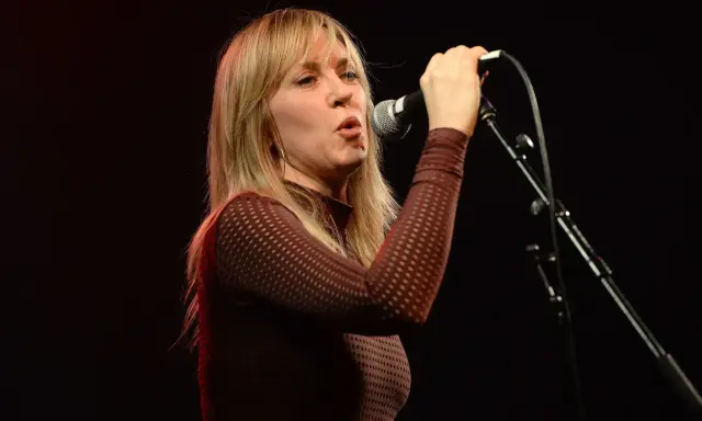 Liz Phair chante avec passion dans un micro, ses cheveux blonds encadrant son visage expressif.