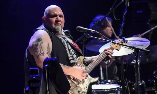 Un guitariste popa chubby, chauve, tatoué, joue sur scène avec un batteur en arrière-plan.