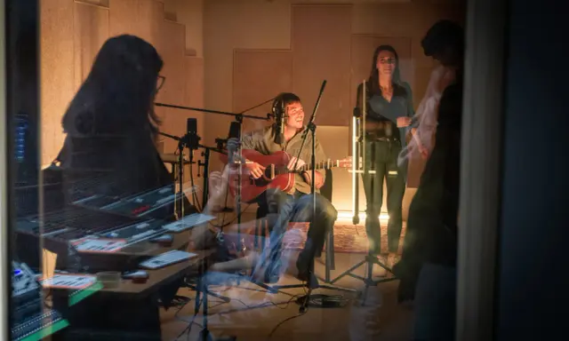 Un homme joue de la guitare dans un studio d'enregistrement, entouré de microphones. Une femme chante à ses côtés. La scène suggère une collaboration musicale, peut-être pour une liste producteur de musique.
