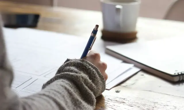 Main tenant une plume bleue, une personne écrit sur du papier, une tasse de café à proximité. Une idée de chanson à écrire prend forme.