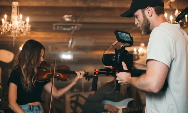 Un caméraman s'apprête à créer un clip vidéo, filmant une violoniste et un guitariste dans un décor chaleureux avec des lustres.