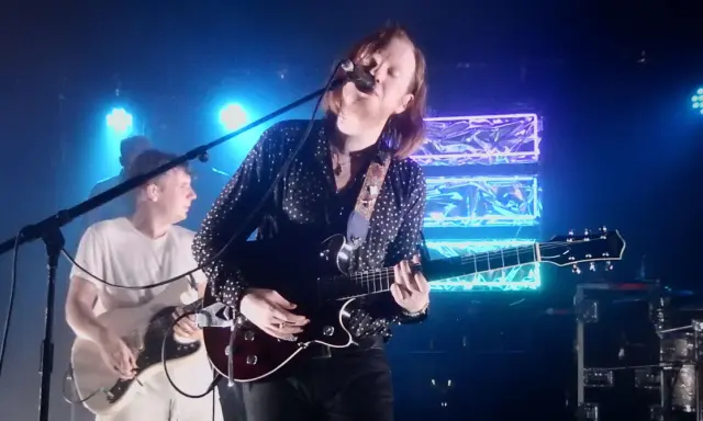 Le groupe Two Doors Cinema Club en concert, éclairé par des néons bleus. Le chanteur joue de la guitare et chante dans un micro.