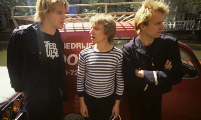 Le groupe Police, dans les années 80, pose devant un véhicule rouge. L'un porte un t-shirt à rayures, les autres des vestes sombres.