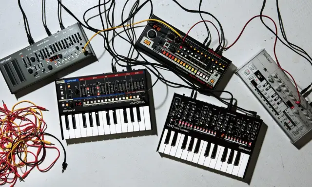 Plusieurs petits instruments de musique électroniques, dont des synthétiseurs et des boîtes à rythmes, sont connectés par des câbles.