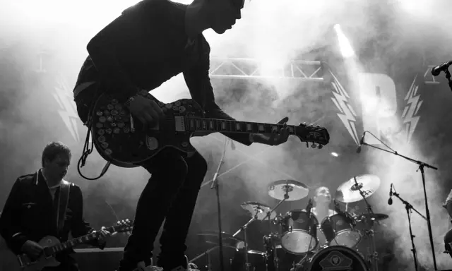 Un guitariste électrique joue sur scène, entouré de fumée. Derrière lui, un batteur et le logo d'un nom de groupe de rock.