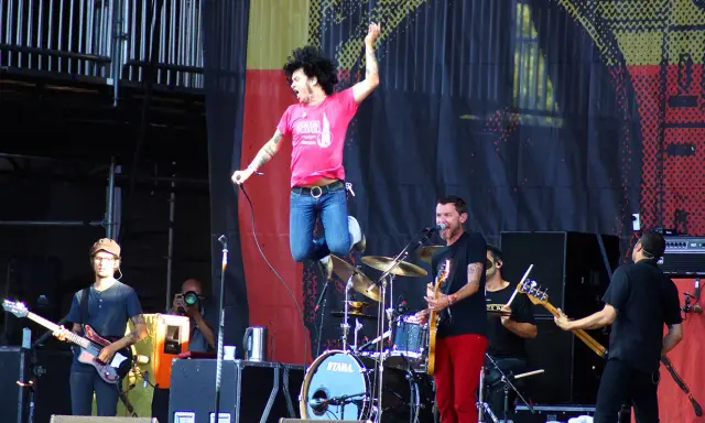 Un chanteur saute sur scène, micro en main, lors d'un concert en plein air. Le groupe joue avec énergie, une ambiance de festival.