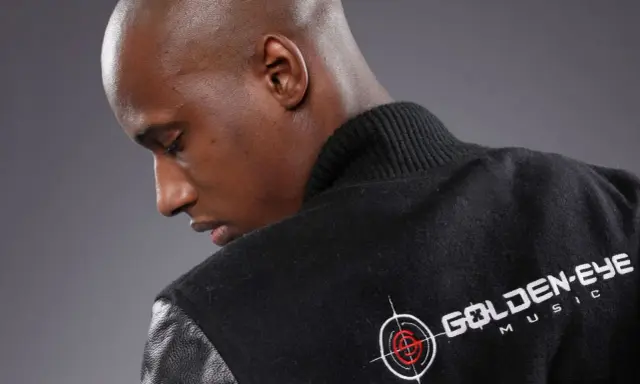 Un homme chauve porte une veste noire avec le logo "Golden Eye Music". Il est prêt à faire le buzz avec sa musique.