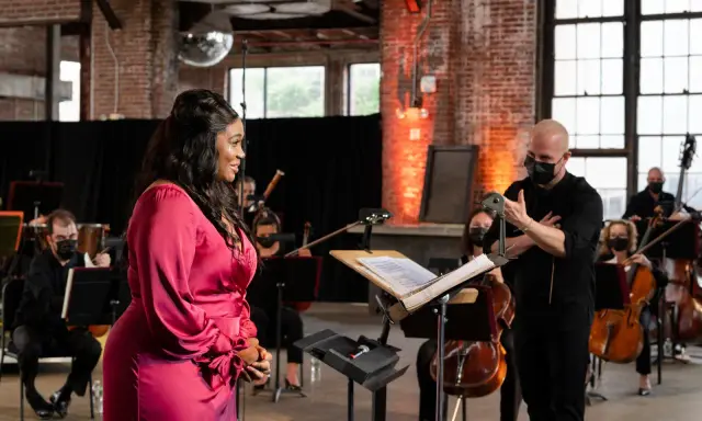 Une chanteuse en robe rose discute avec un chef d'orchestre masqué, entourée de musiciens. Elle cherche peut-être à trouver un agent pour sa carrière.