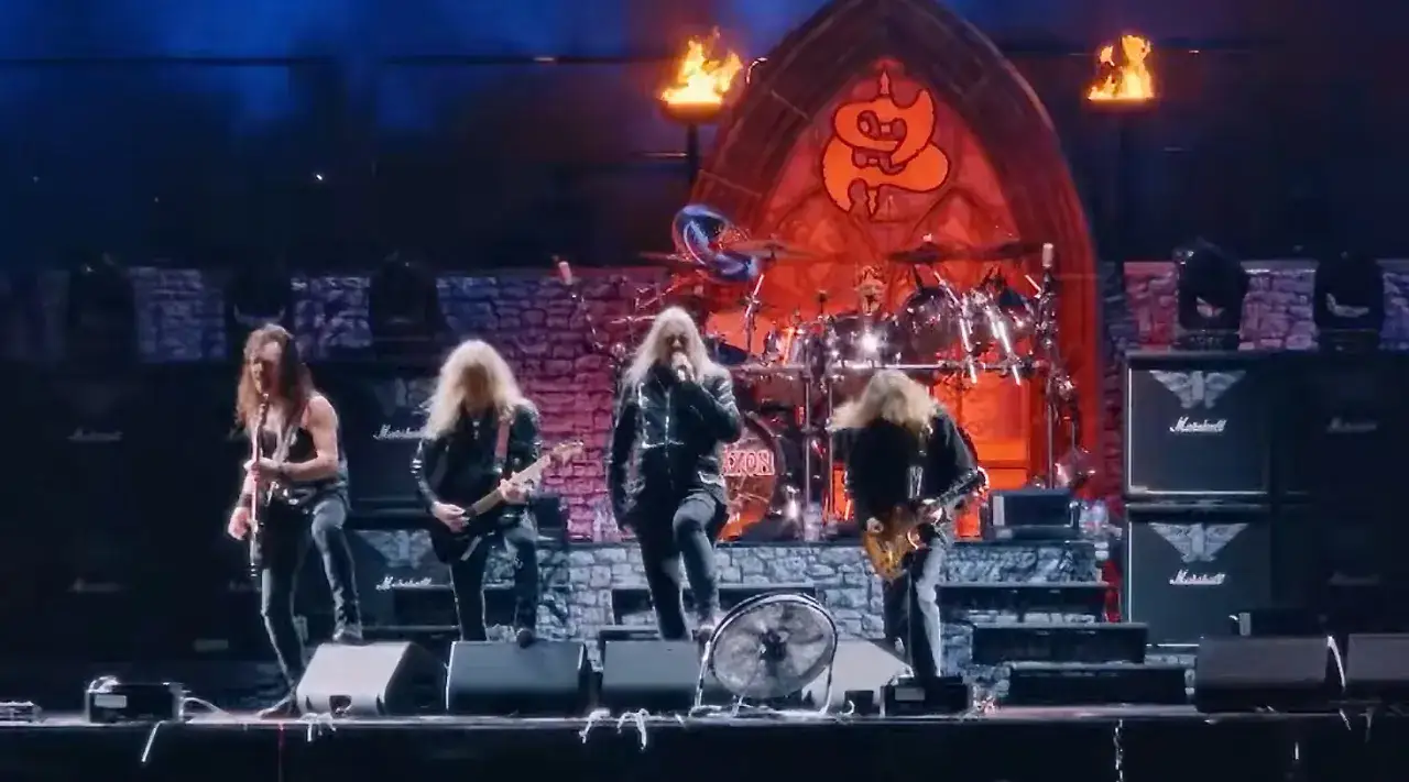 Le groupe Saxon en pleine performance, avec des guitares, une batterie et un chanteur charismatique sur sc&egrave;ne.