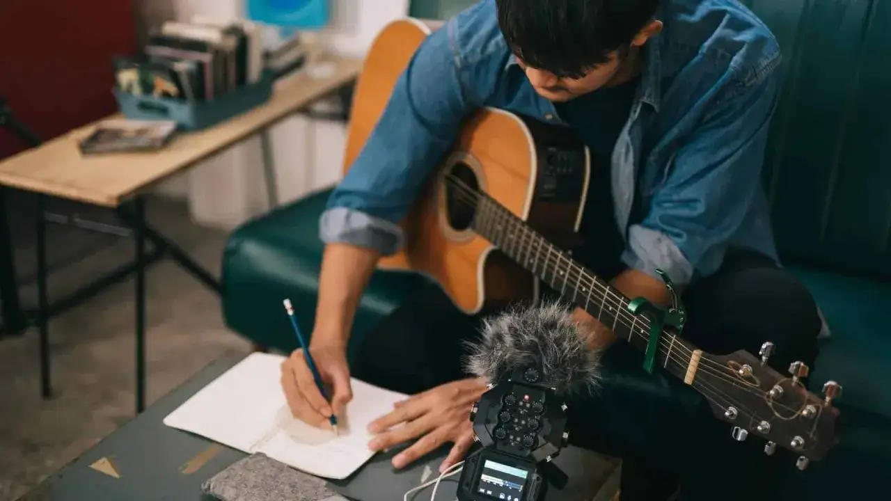Un jeune homme avec une guitare cherche comment trouver l'inspiration pour écrire une chanson, notant ses idées dans un carnet.