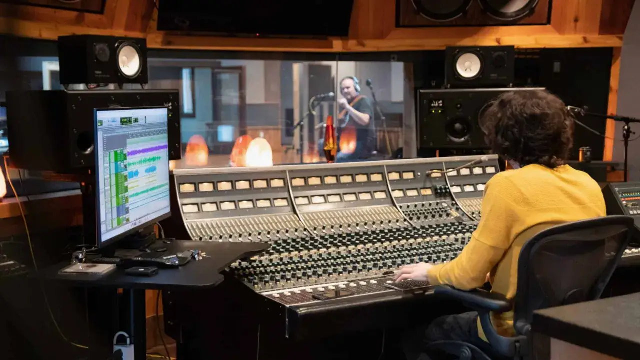 Ing&eacute;nieur du son ajustant les r&eacute;glages sur une console pour cr&eacute;er un effet de reverb definition musique, avec un musicien en studio derri&egrave;re la vitre.