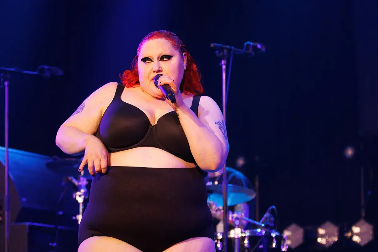Une chanteuse aux cheveux rouges, maquillage audacieux, porte un soutien-gorge noir et une culotte haute sur scène. Elle tient un micro, prête à partager ses histoires avec le gossip groupe.
