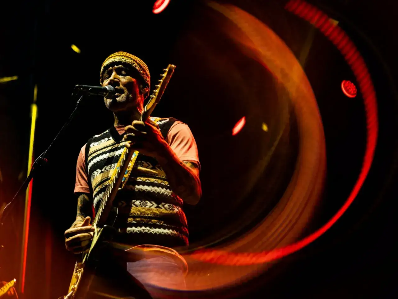 Ben Harper, le musicien, joue de la guitare sur scène, baigné de lumières chaudes et tourbillonnantes.