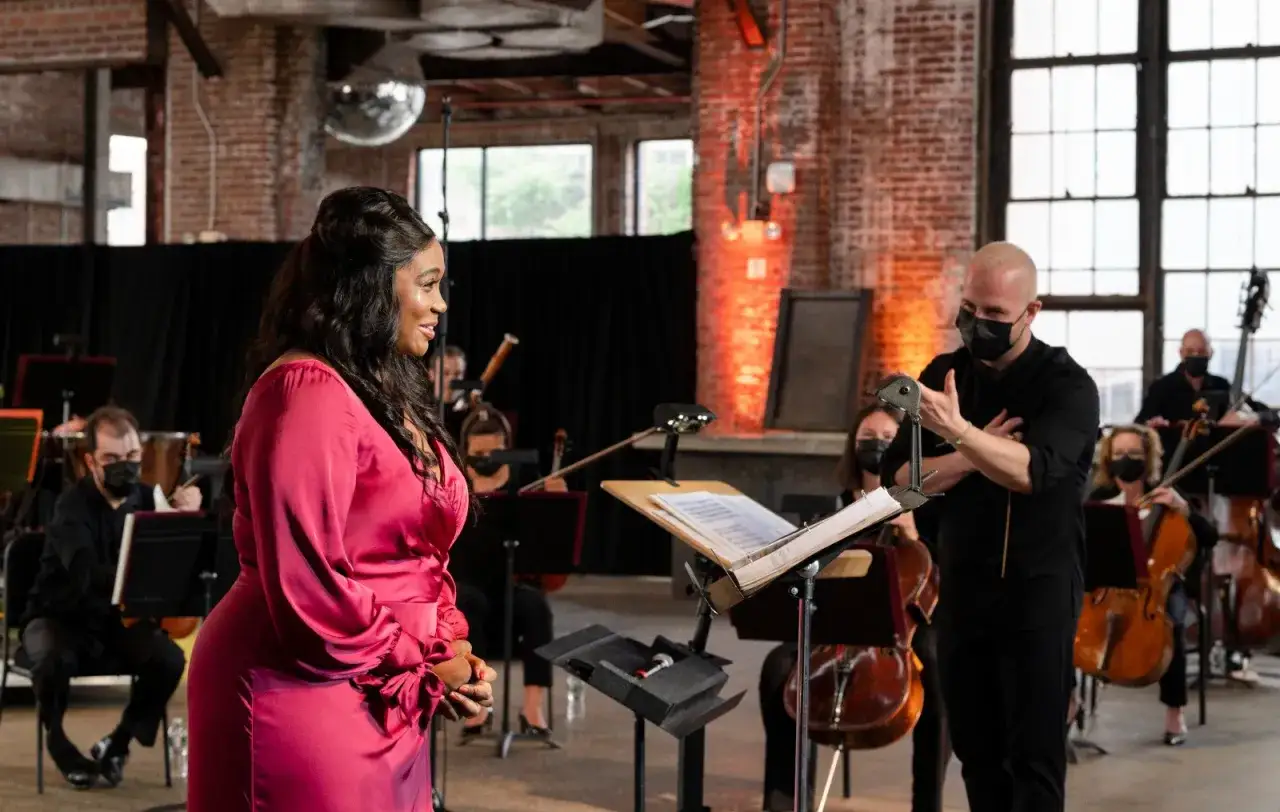 Une chanteuse en robe rose dialogue avec le manager du groupe de musique, un orchestre jouant en arrière-plan.