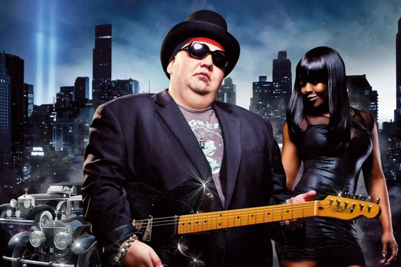 Un homme popa chubby, chapeau noir, lunettes de soleil, joue de la guitare électrique devant une femme en robe noire et un paysage urbain nocturne.