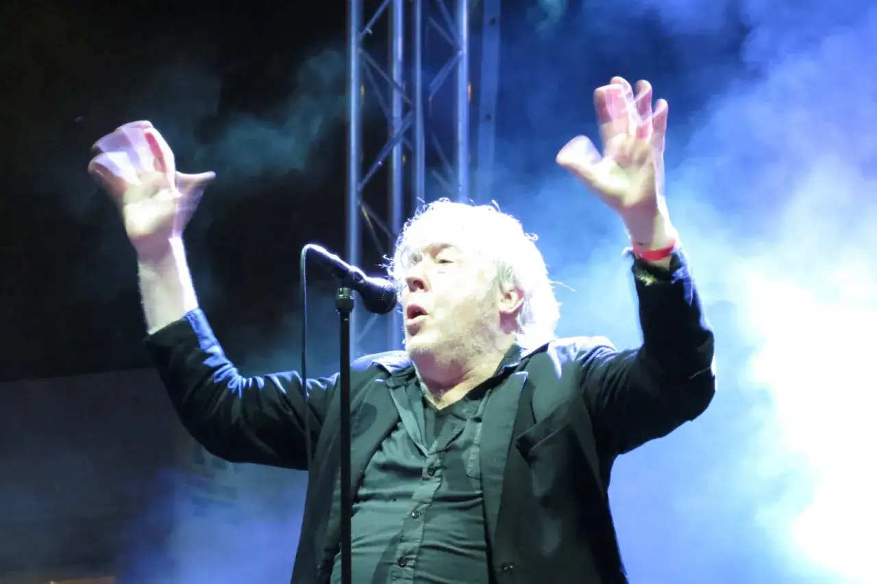 Arno, chanteur emblématique, enflamme la scène, mains levées dans un geste passionné, sous les lumières bleutées.