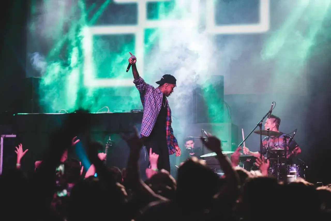 Un rappeur sur scène, micro en main, encourage la foule à promouvoir sa musique. Un batteur l'accompagne, sous des lumières vertes.
