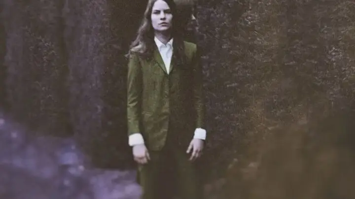 Eliot Sumner, en costume vert, pose devant un mur de pierre sombre.
