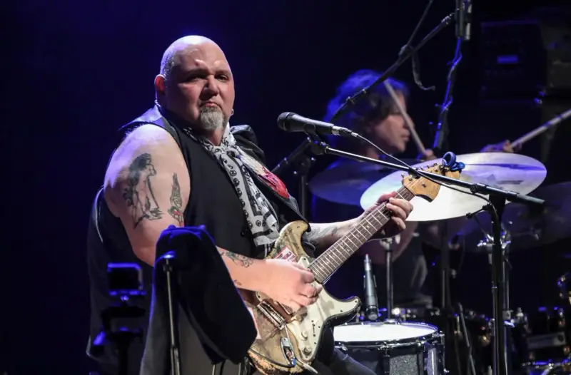 Un guitariste popa chubby, chauve, tatoué, joue sur scène avec un batteur en arrière-plan.