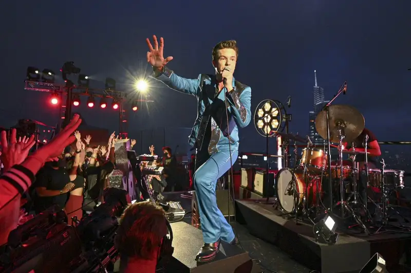 Brandon Flowers de The Killers, en costume bleu scintillant, salue la foule lors d'un concert. Les albums de The Killers résonnent dans la nuit.